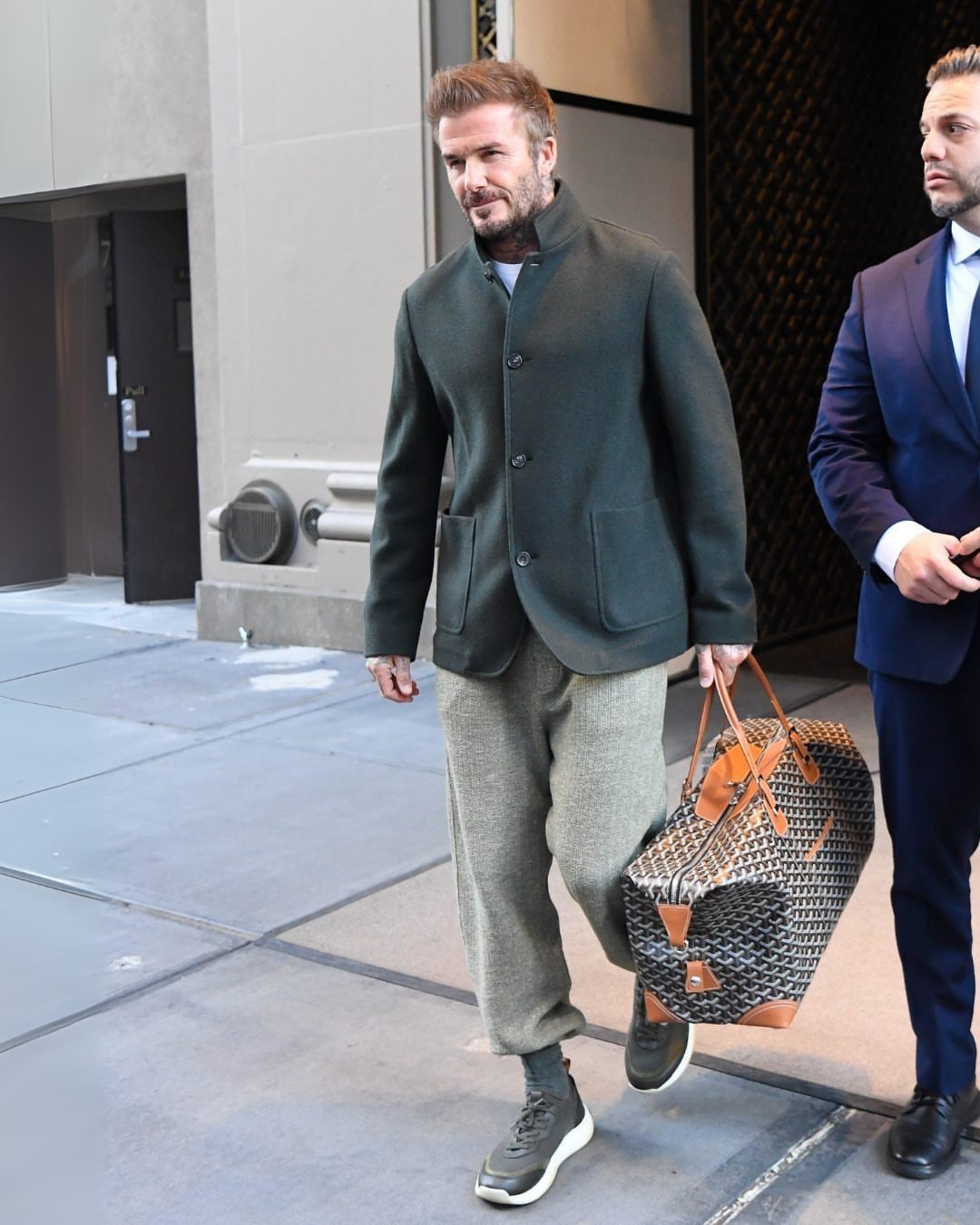 David Beckham walking in a dark green jacket and grey trousers carrying a large patterned bag.