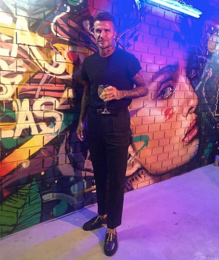 David Beckham posing in a black t-shirt and trousers wearing black loafers against graffiti.