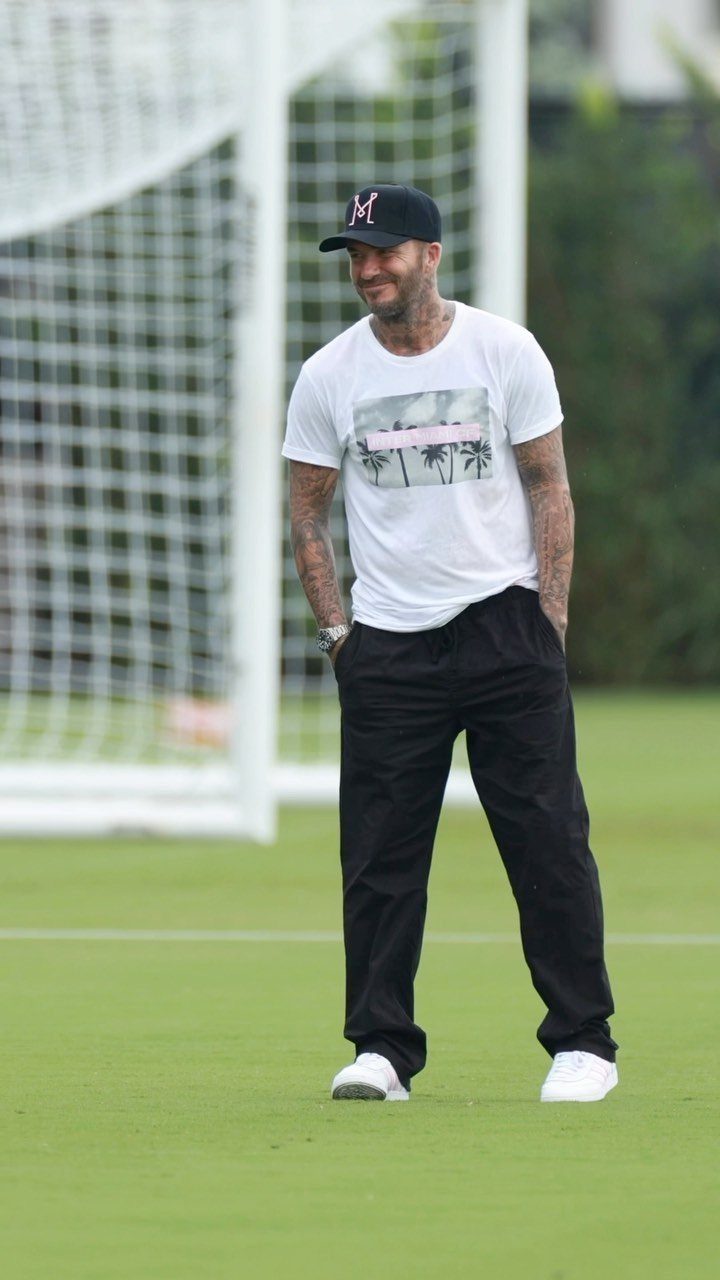 David Beckham wearing a white graphic t-shirt, black trousers, and a black cap on a soccer field.