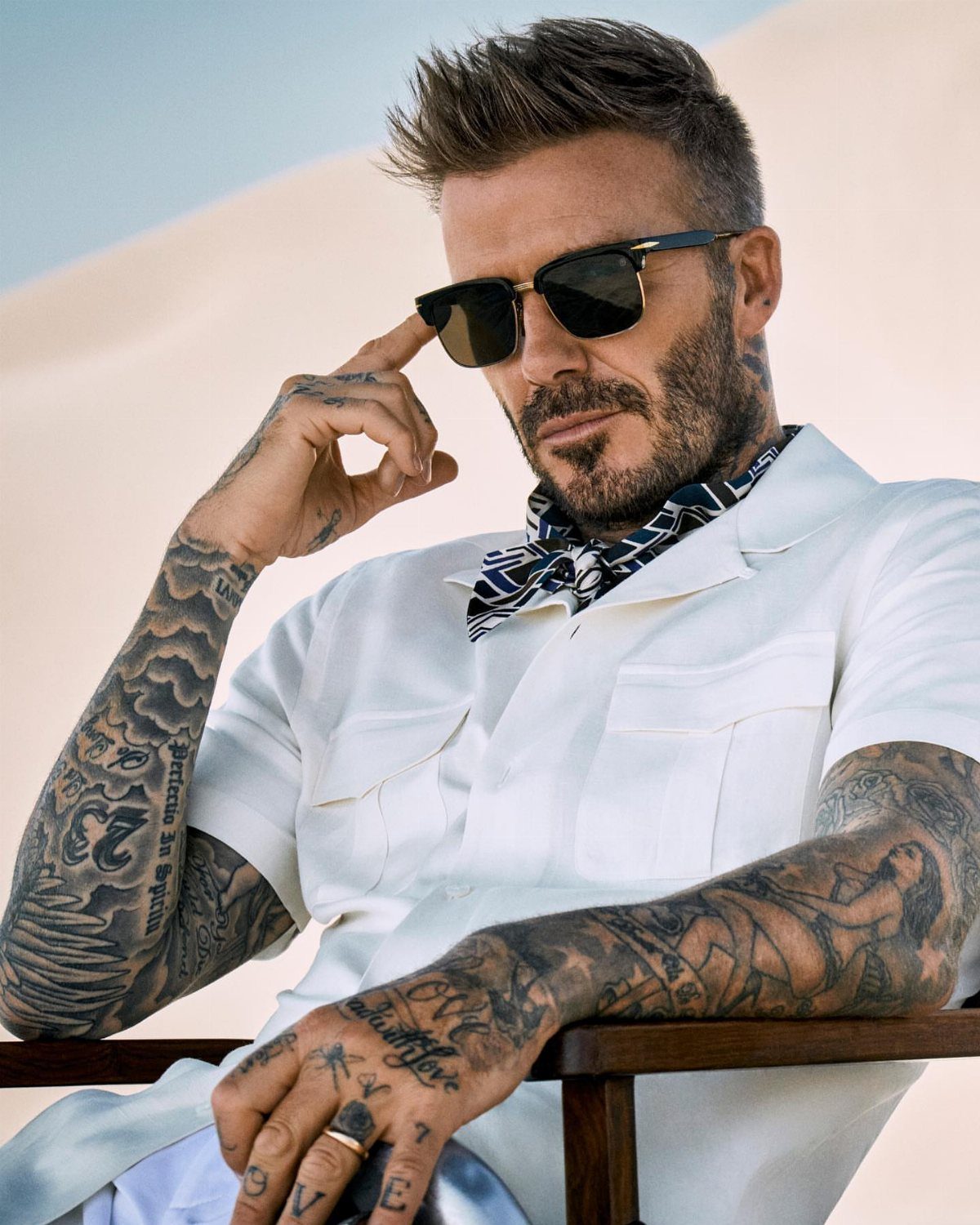 A man with spiky hair and sunglasses wearing a white shirt and patterned neck scarf.