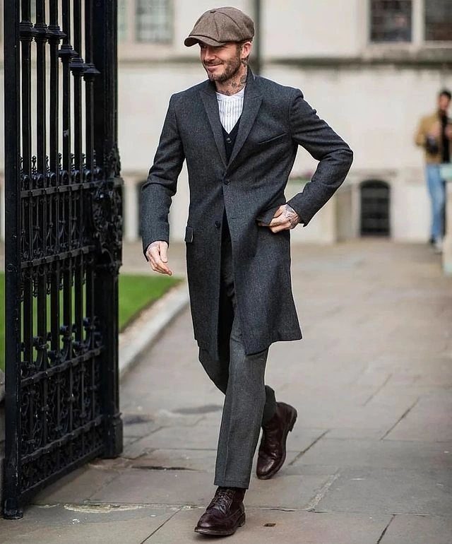 A man walking in a long charcoal grey coat, trousers, and a brown flat cap.