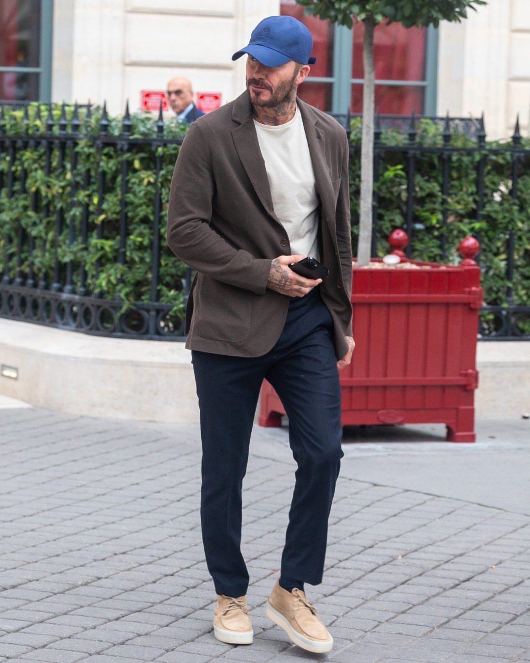 Man in a brown blazer, white t-shirt, navy trousers, and blue cap walking outdoors.