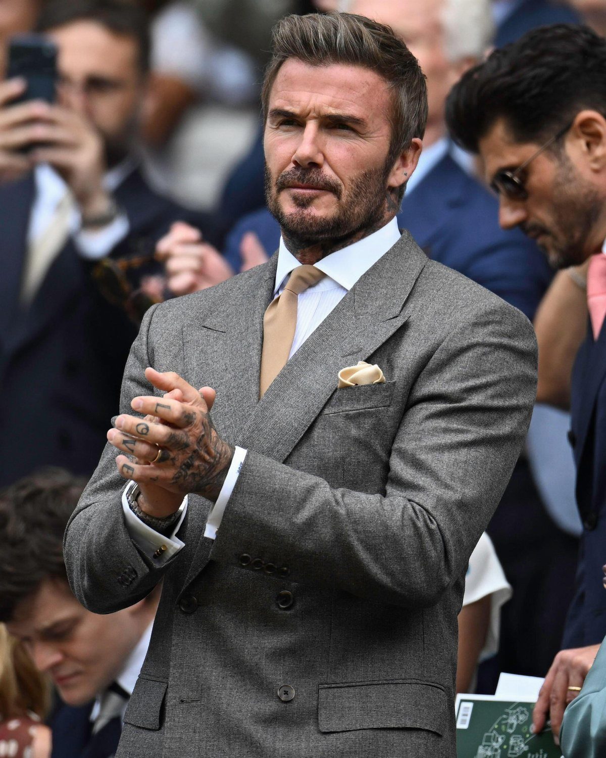 David Beckham in a grey patterned double-breasted suit and tan tie, clapping his hands.