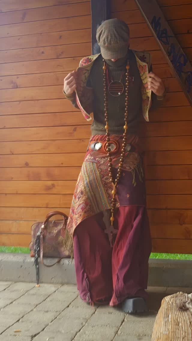 A person wearing maroon flowy pants, patterned vest, and long beaded necklaces against a wooden wall.