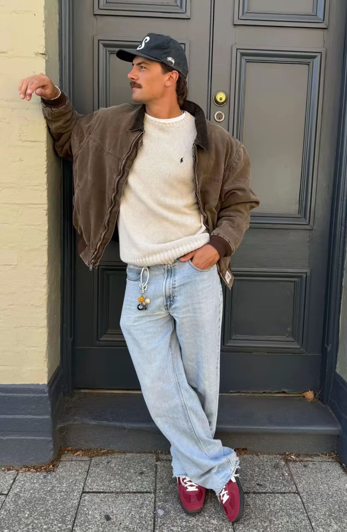 A man in a brown suede jacket, white sweater, and light blue jeans leaning against a wall.
