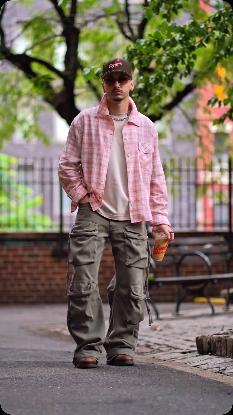 A man in a pink plaid shirt and olive cargo pants walking through a park.