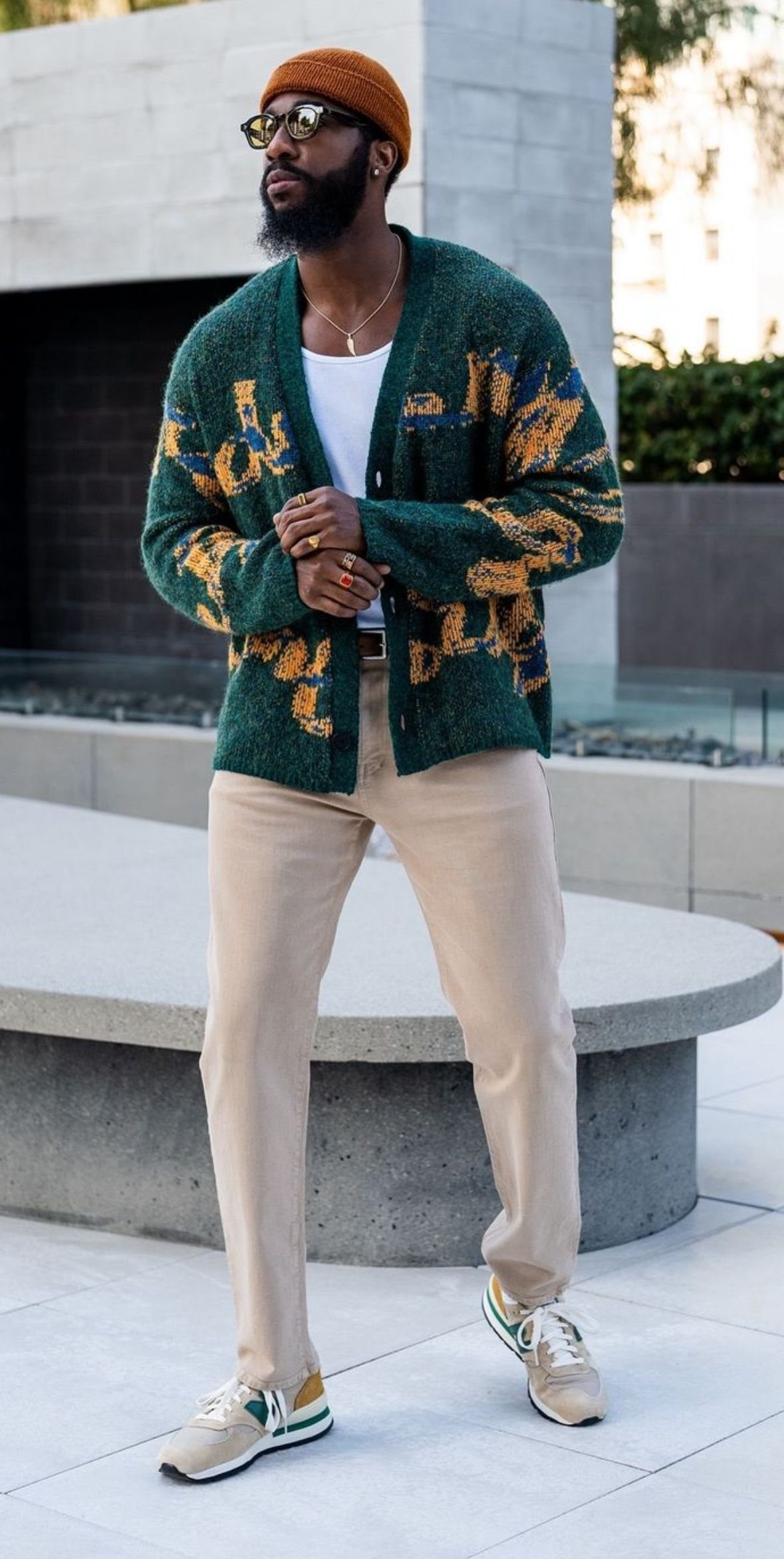A man wearing a bold patterned green cardigan, beige pants, and a brown beanie.