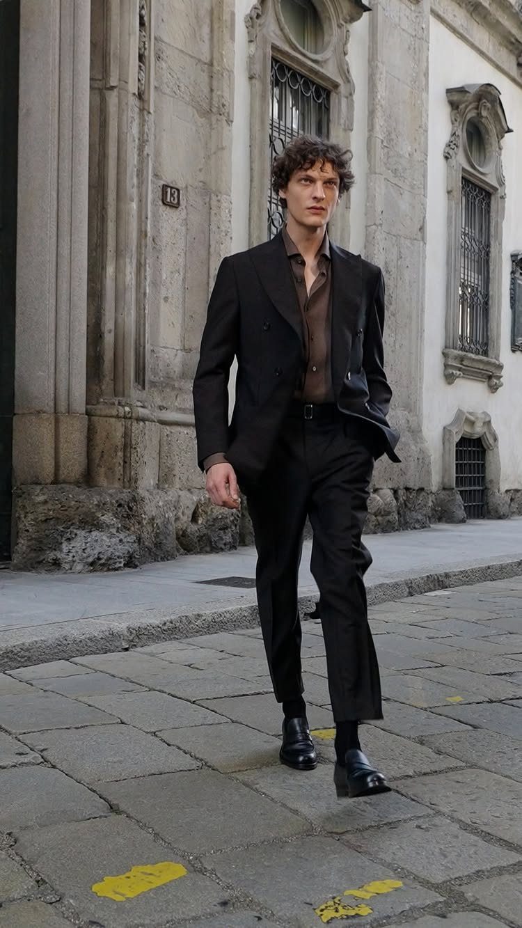 A man walking down a city street wearing a dark double-breasted suit.
