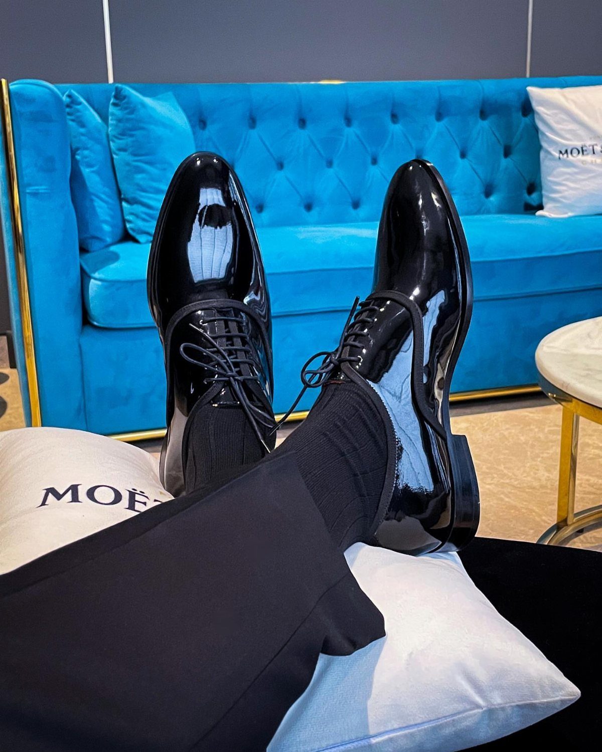 Polished black patent leather dress shoes worn with black trousers and socks against a blue sofa.