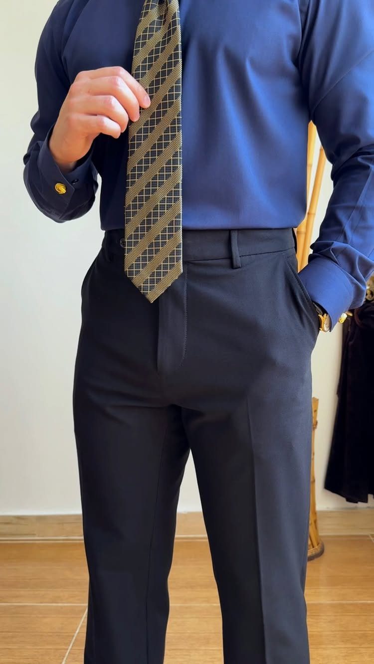A man in a blue shirt and dark trousers holding a patterned gold tie.