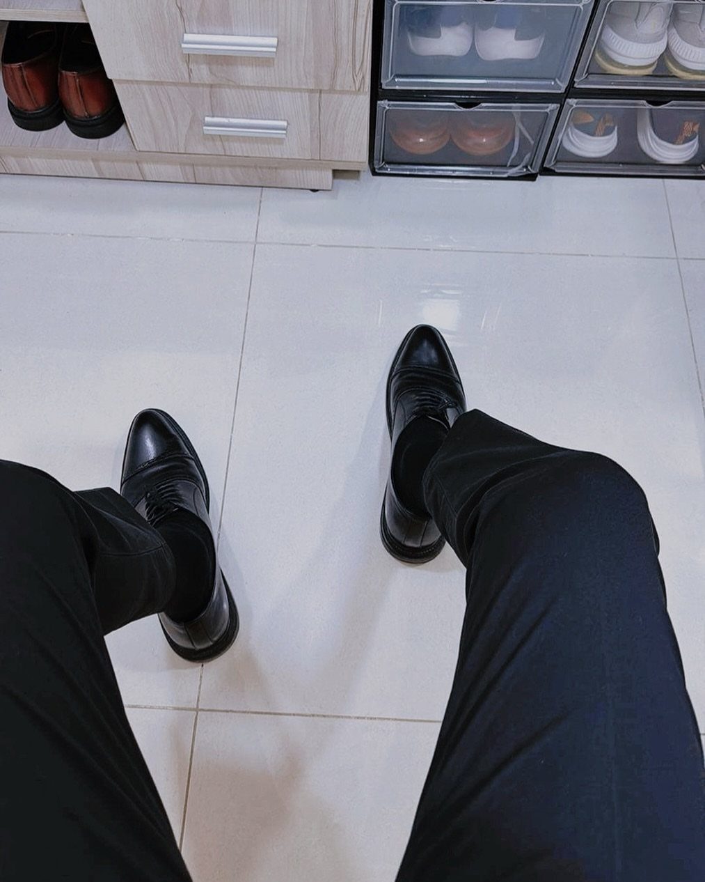 Black leather oxford shoes worn with black formal trousers on a light tiled floor.