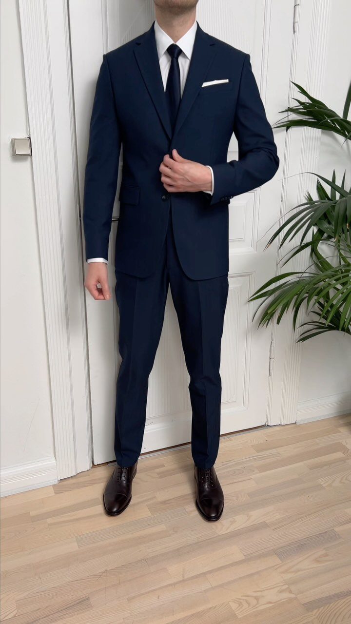 A man wearing a navy blue suit, white shirt, and navy tie standing indoors.