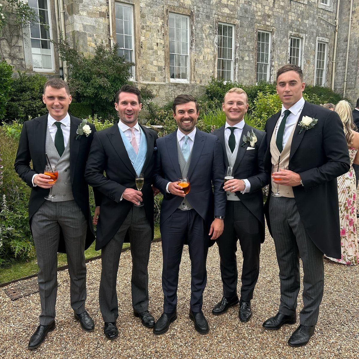 Five men smiling while posing in formal wedding attire in front of a stone building.
