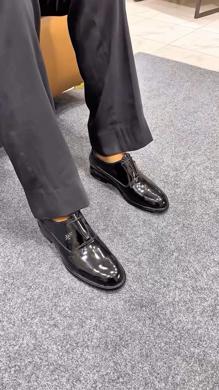 Black patent leather dress shoes worn with black formal trousers on a grey carpet.