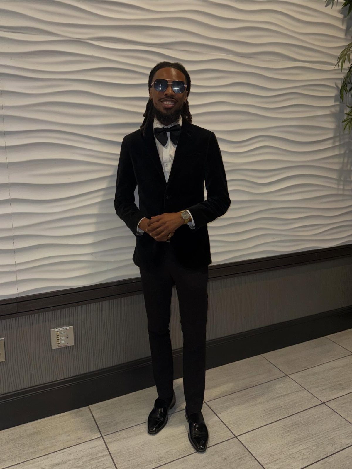 Man in a black velvet tuxedo and bowtie posing against a white wavy wall.