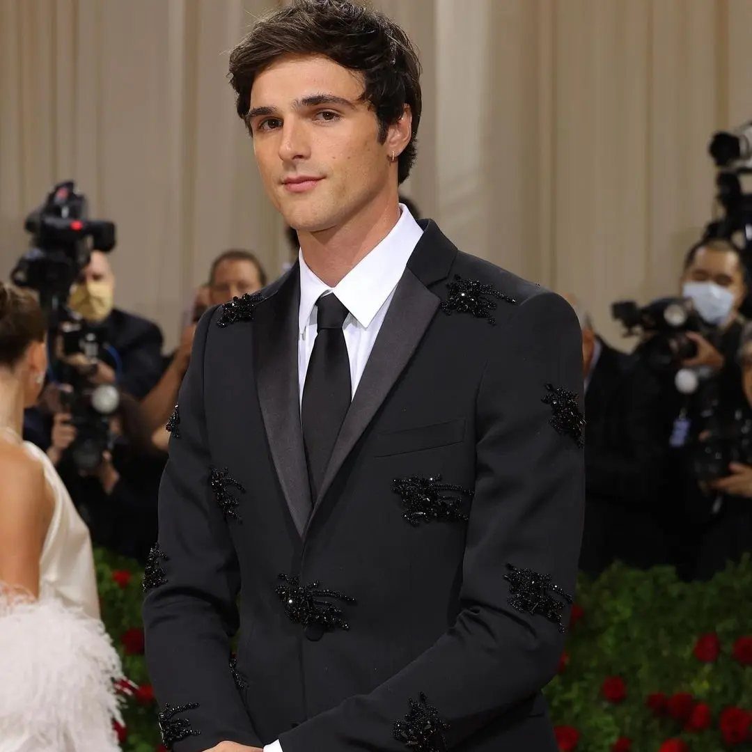 A man wearing a black embellished tuxedo jacket, white shirt, and black tie.