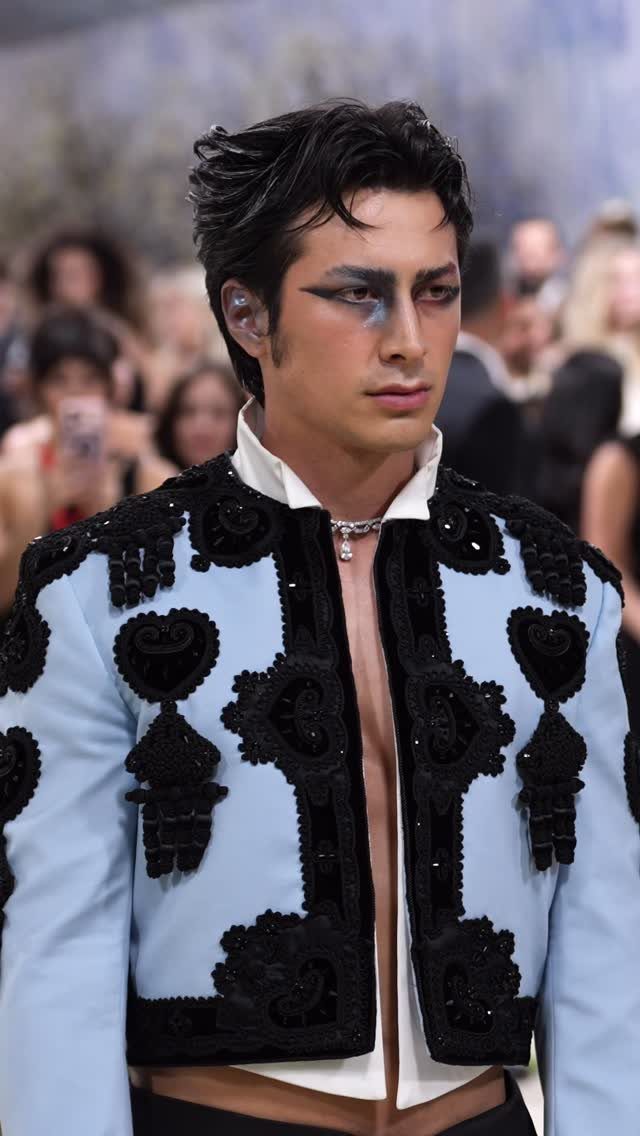 Man with dramatic eye makeup wearing an ornate light blue and black embroidered jacket.