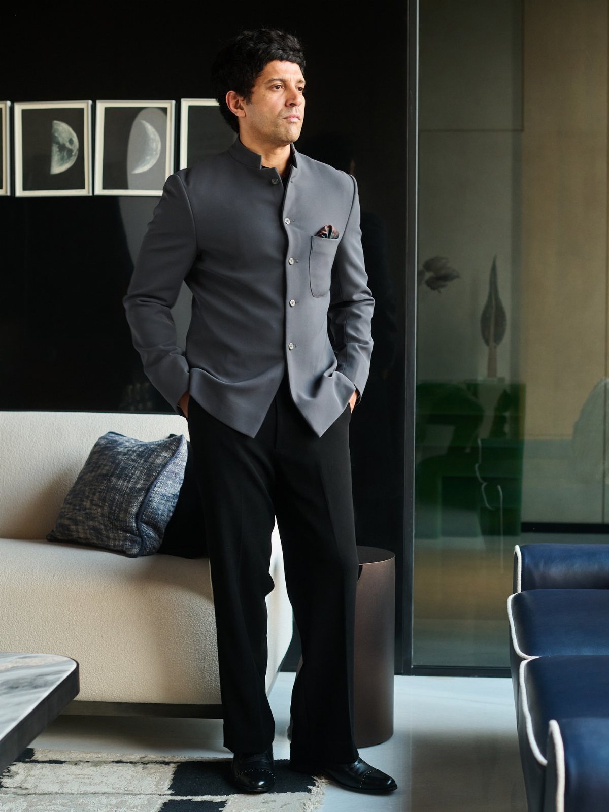 A man posing in a grey Nehru jacket, black trousers, and black dress shoes in a modern room.