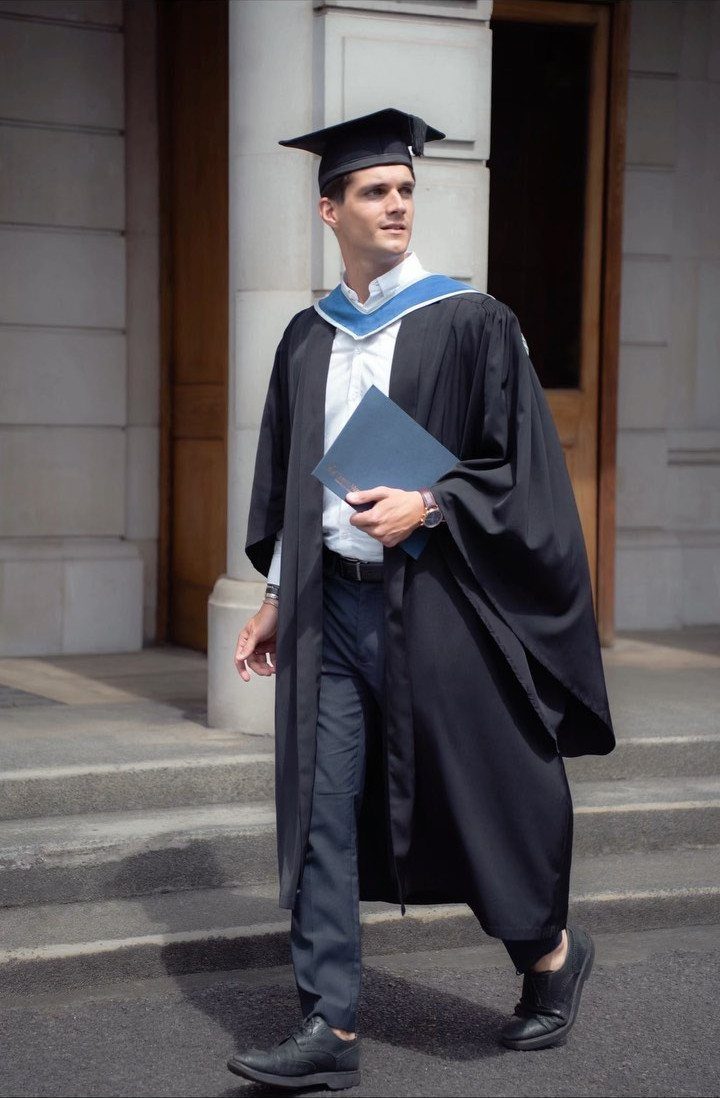 A man in a black graduation gown and cap walking while holding a blue folder.