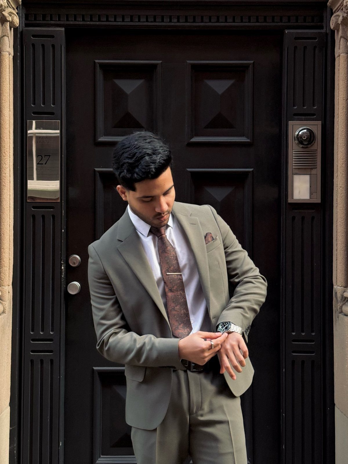 Man in a grey suit and patterned tie adjusting his watch in front of a dark door.