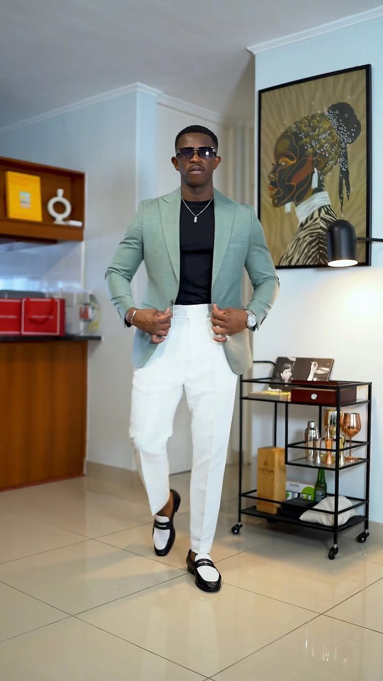 Man wearing a light green blazer, black shirt, white pants, and black loafers posing indoors.