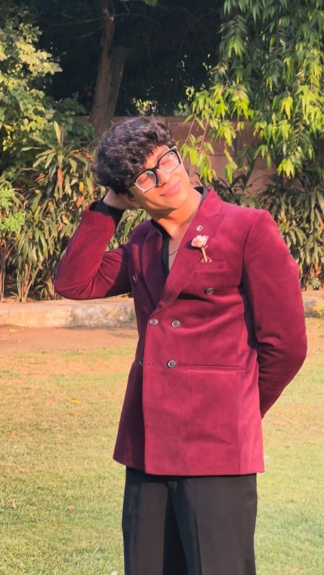 A young man in a maroon velvet blazer and black trousers posing in a garden.