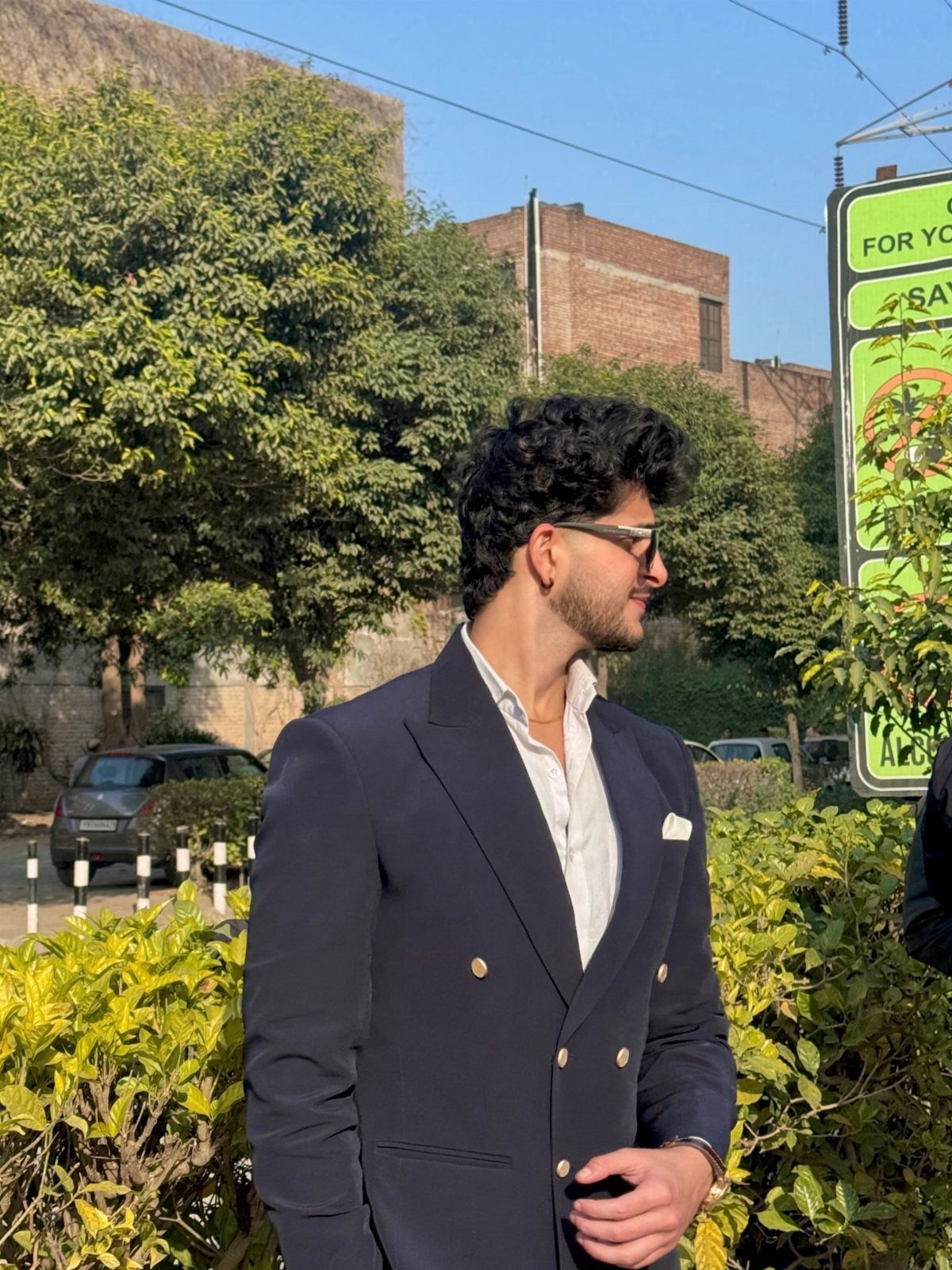 Man in a navy double-breasted blazer and white shirt standing near green bushes outdoors.