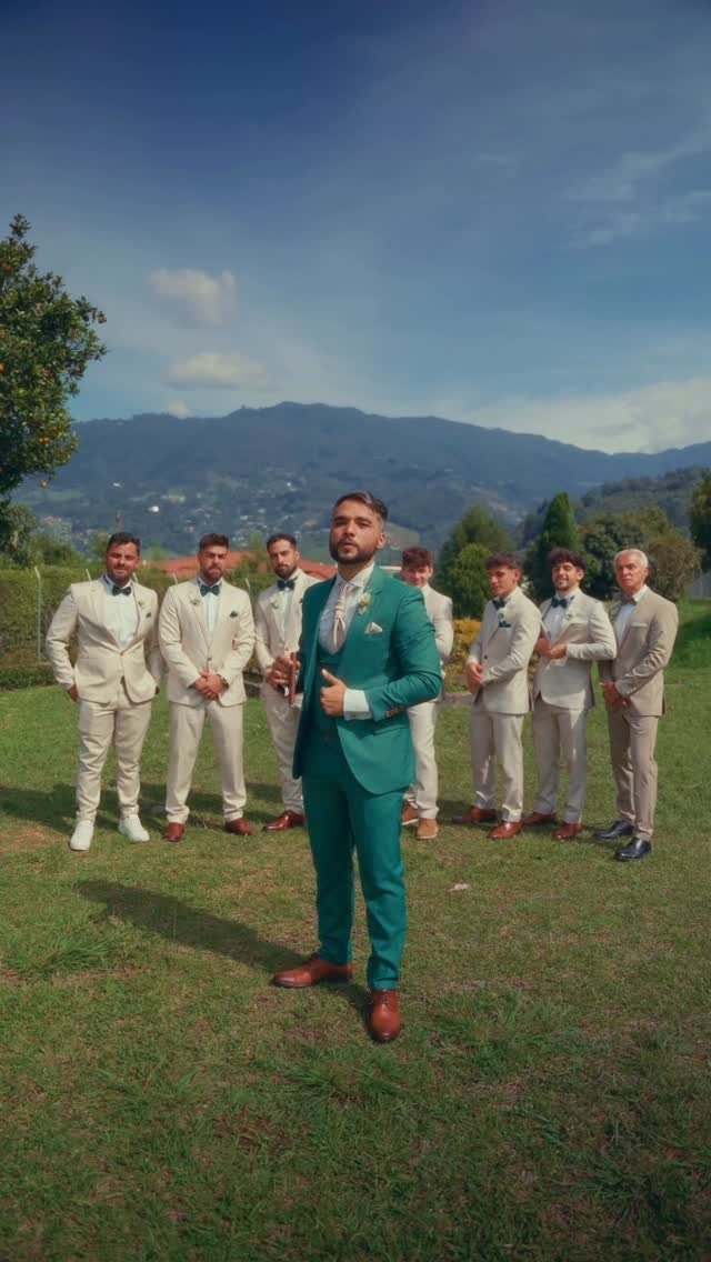 A man in a teal suit stands in front of a group of men wearing beige suits in a grassy field.