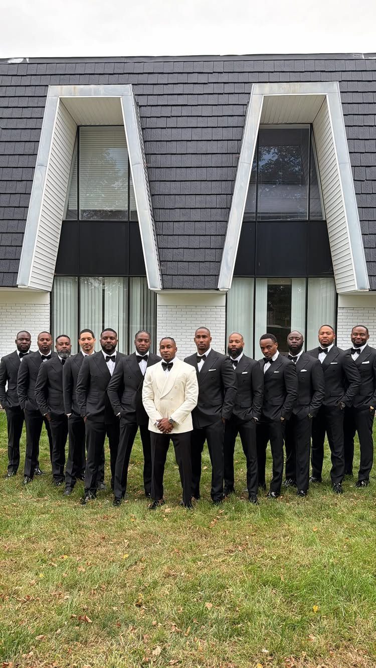 A large group of men posing in matching black and white formal suits outdoors.