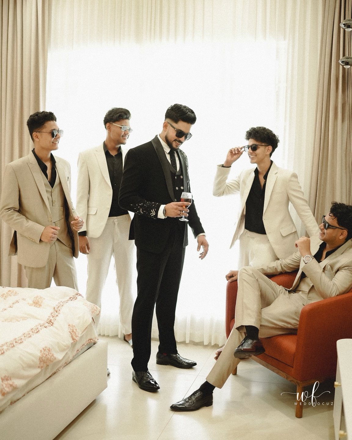 Six men posing in stylish black tuxedos and beige suits while wearing sunglasses indoors.