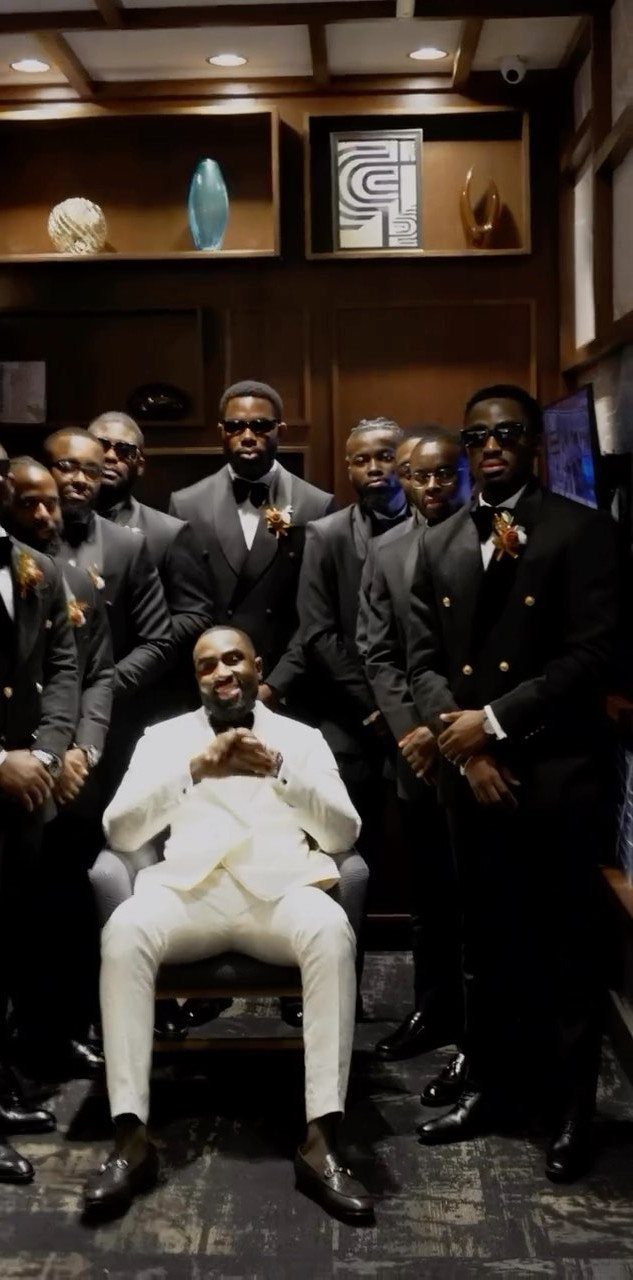 A man in a white suit sits surrounded by groomsmen in black suits.