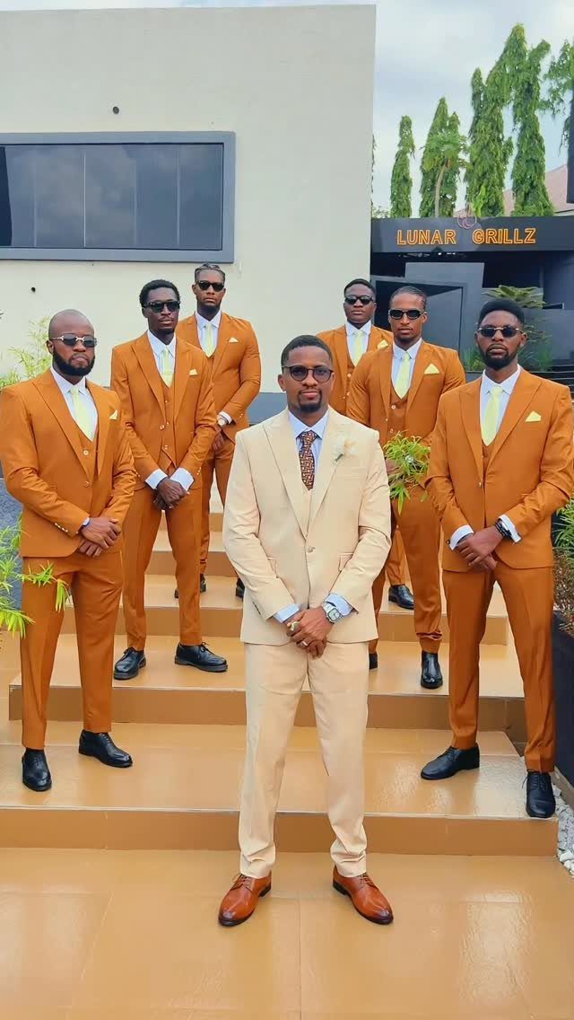 Seven men posing in mustard yellow and beige formal suits on outdoor stairs.