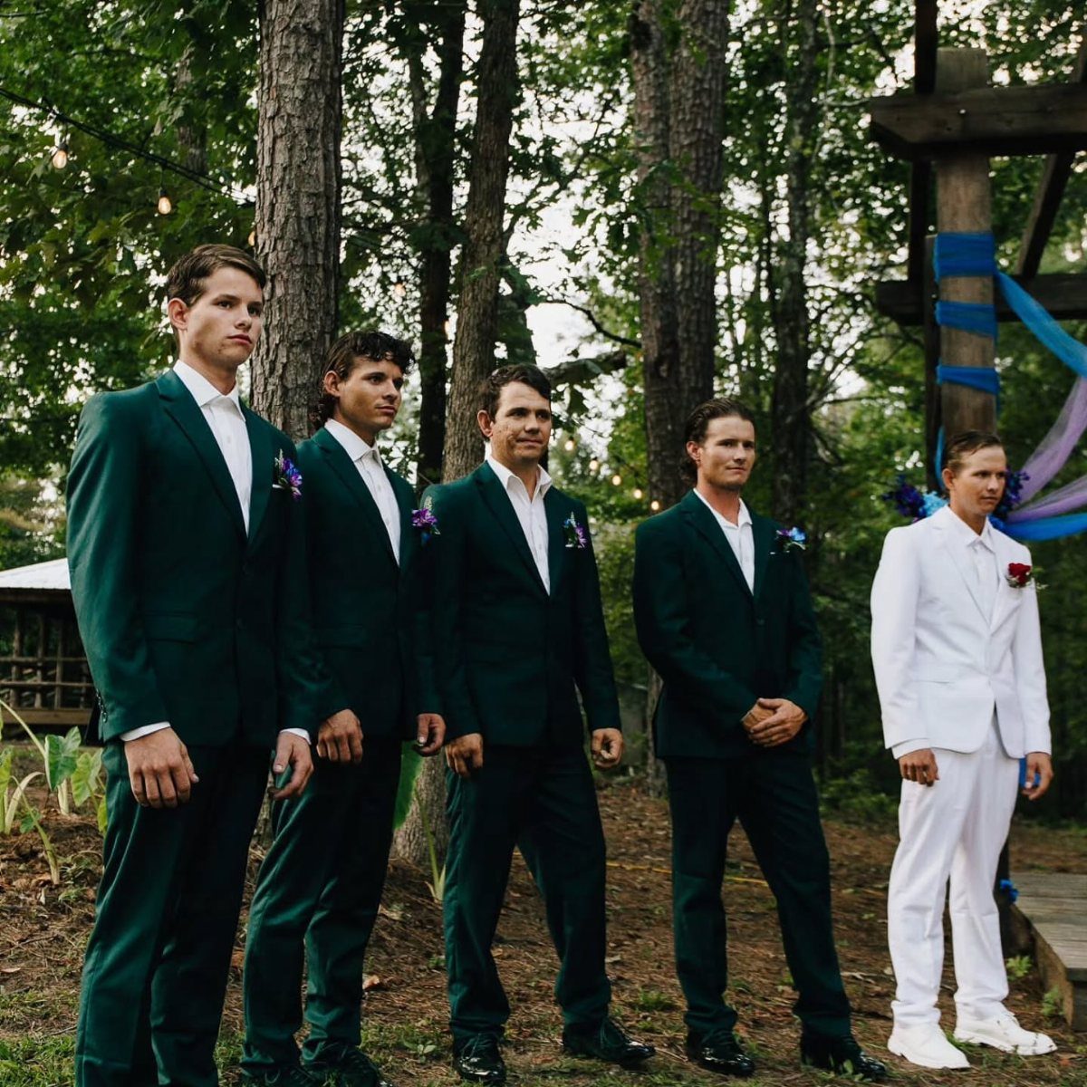 Five men in formal suits standing in a row in a wooded outdoor setting.