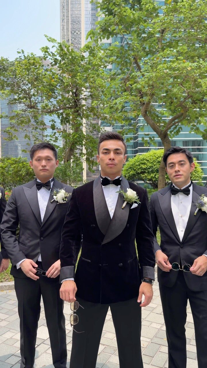 Three men in formal black tuxedos and bow ties standing outdoors near green trees and skyscrapers.