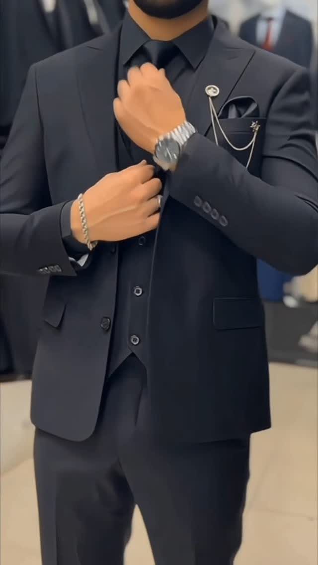 Man in a black three-piece suit adjusting his tie and wearing silver accessories.