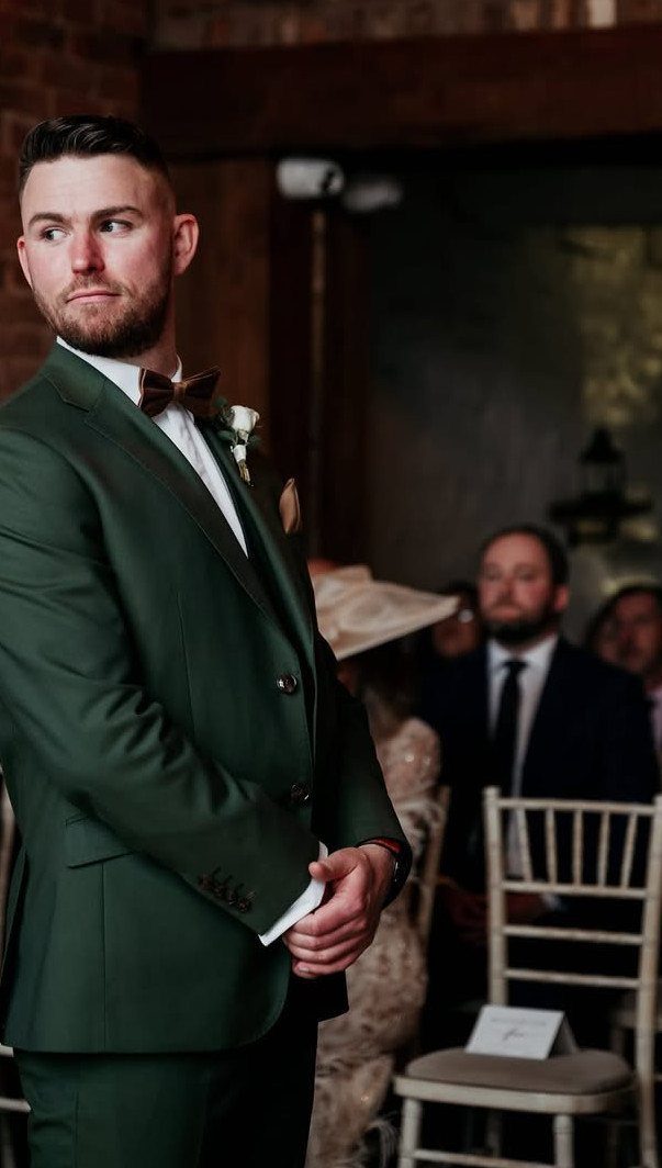 Man in a dark green suit and brown bowtie looking away during a wedding ceremony.