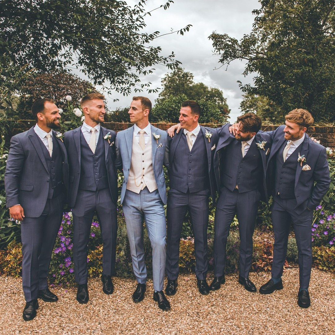 Six men in blue wedding suits posing together in a lush green garden.