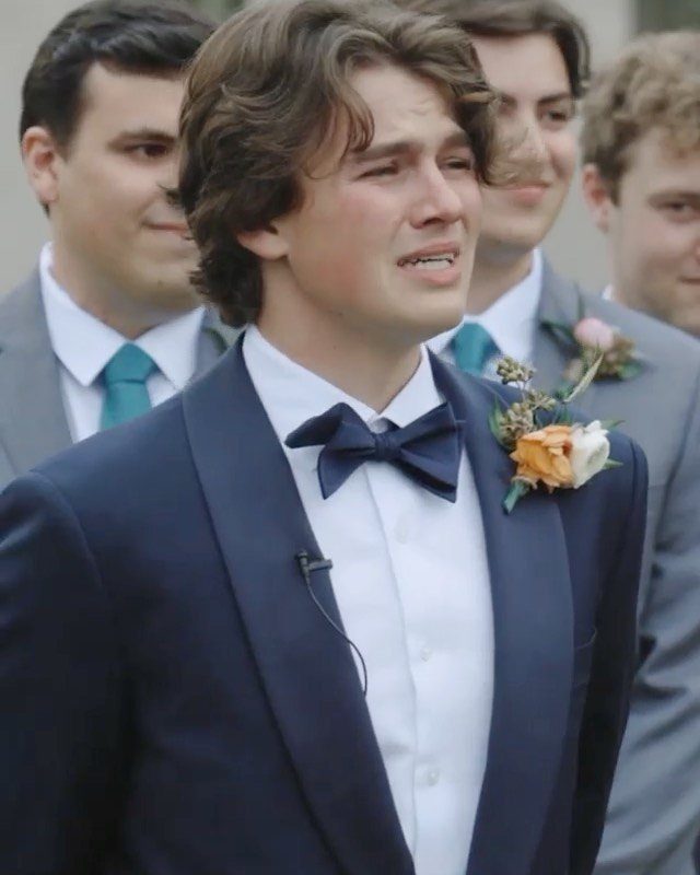 A young man in a navy blue tuxedo and bow tie looking emotional during a wedding ceremony.