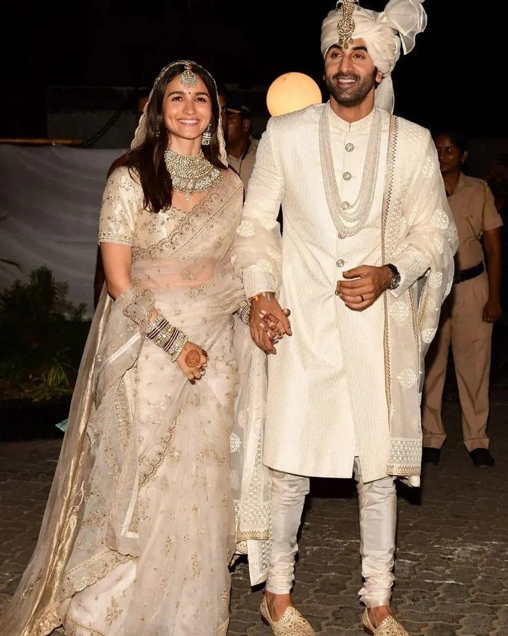 A man and woman smiling while wearing ornate ivory traditional Indian wedding clothing.