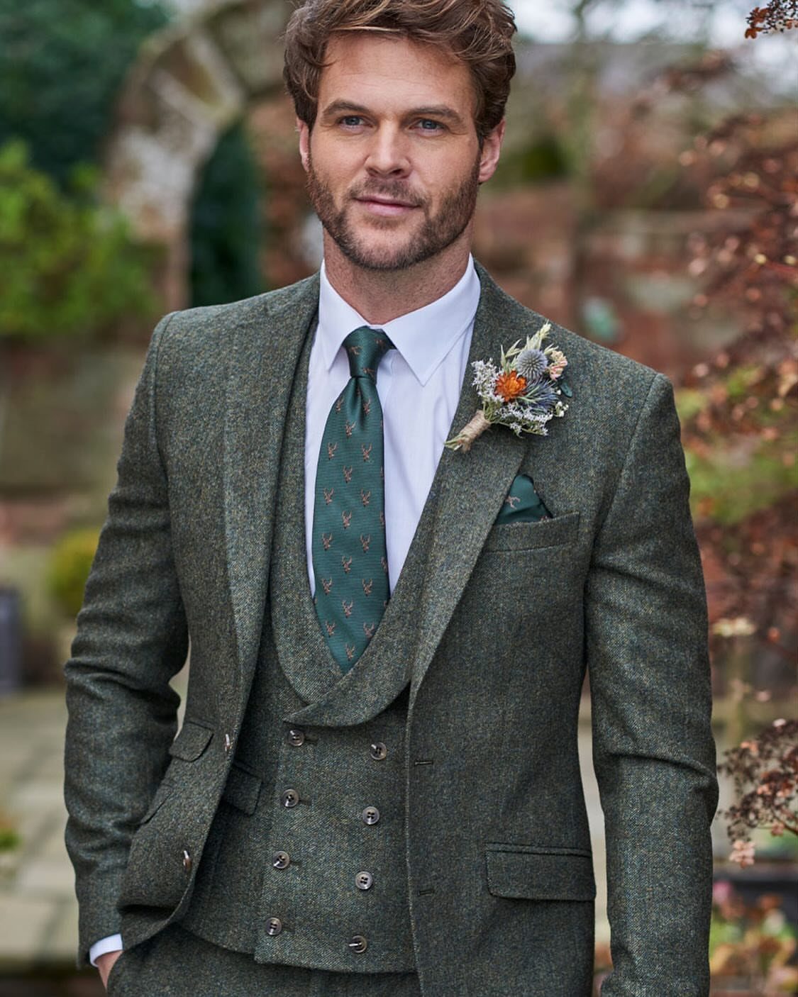 A man posing in a green three-piece tweed suit with a patterned tie and boutonniere.