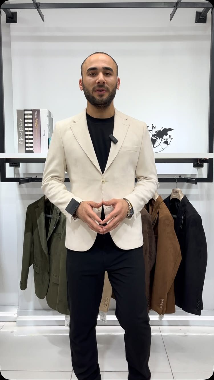 A man wearing a cream blazer, black t-shirt, and black trousers standing in a clothing store.