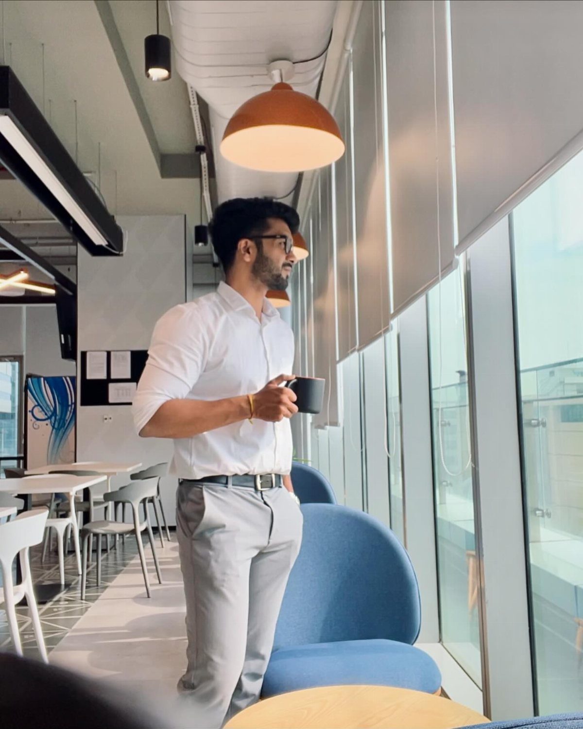 A man in business casual attire stands by an office window holding a coffee mug.