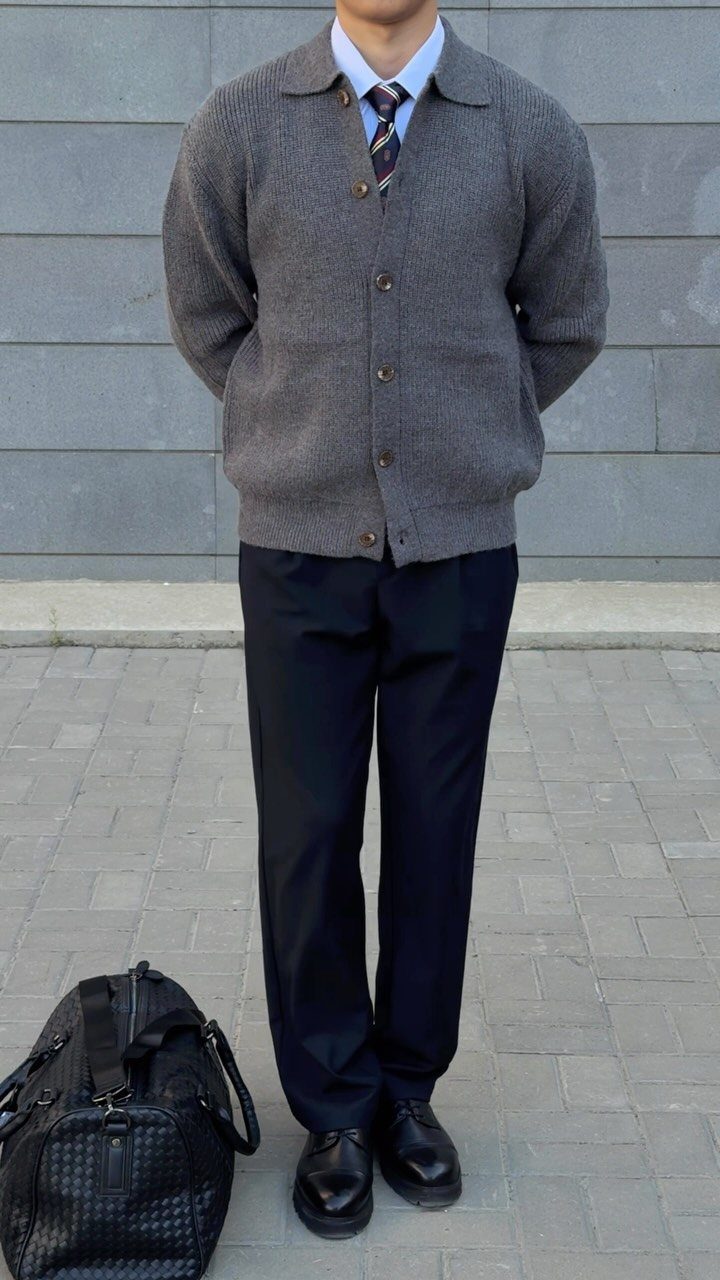 Man wearing a grey cardigan, white shirt, tie, navy trousers, and black shoes standing outdoors.