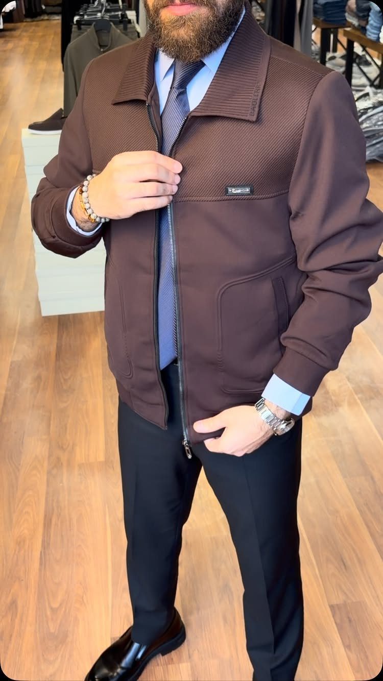 Man wearing a brown textured jacket, blue shirt, tie, and black trousers standing on a wooden floor.
