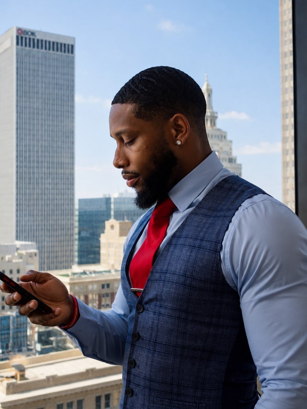 A man in a blue waistcoat and red tie looks at his smartphone against a city skyline.