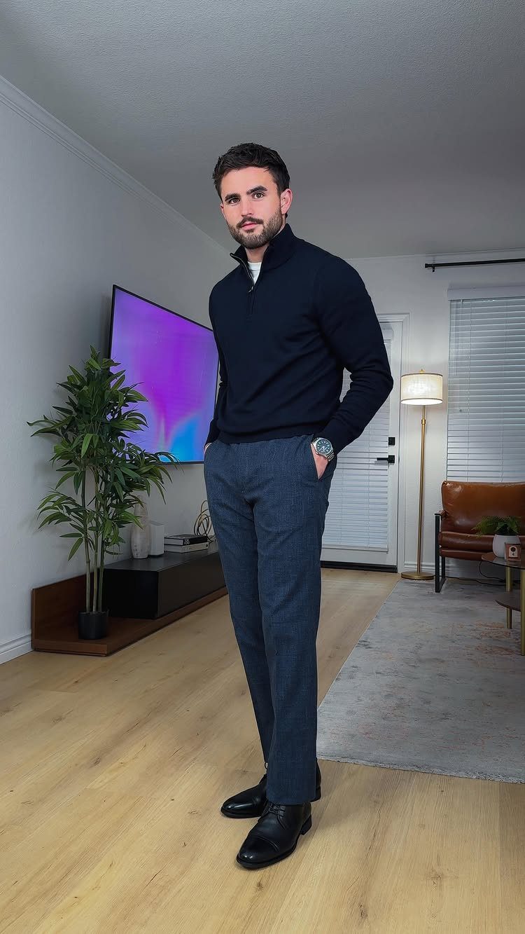 A man posing in a navy quarter-zip sweater, dark trousers, and black leather shoes.
