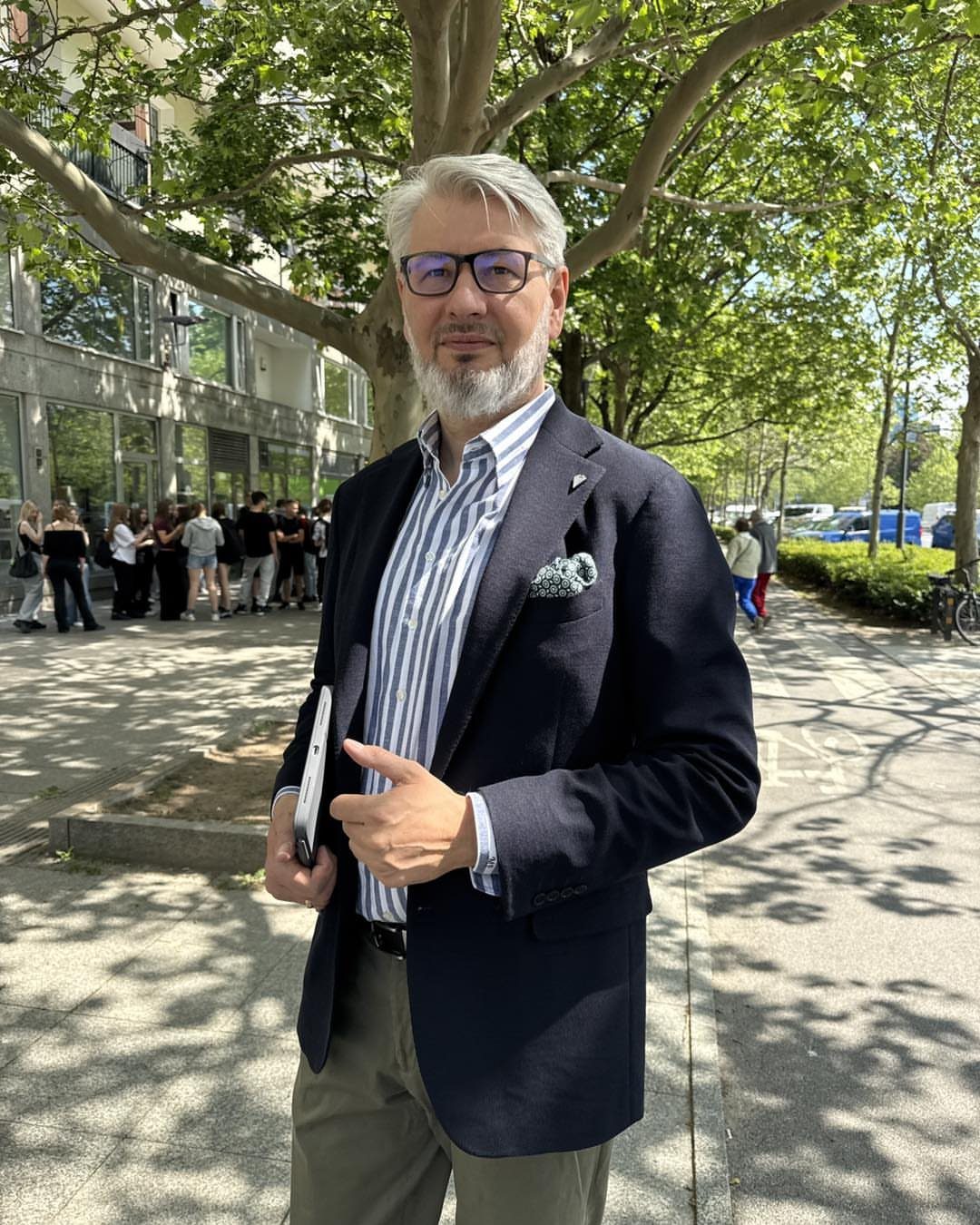 Man with a grey beard wearing a navy blazer and striped shirt standing outdoors.