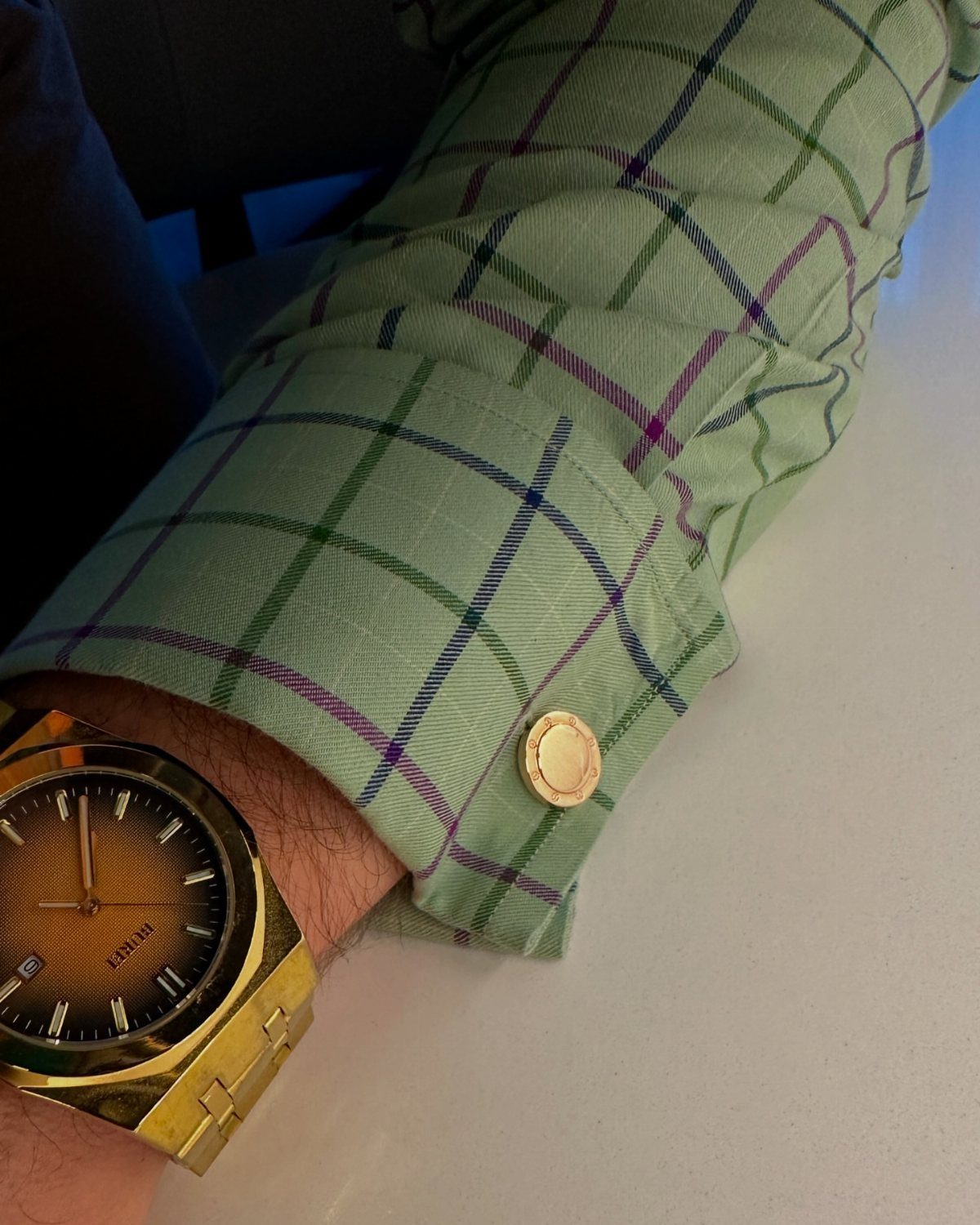 A person wearing a green plaid shirt with a gold cufflink and a gold watch.