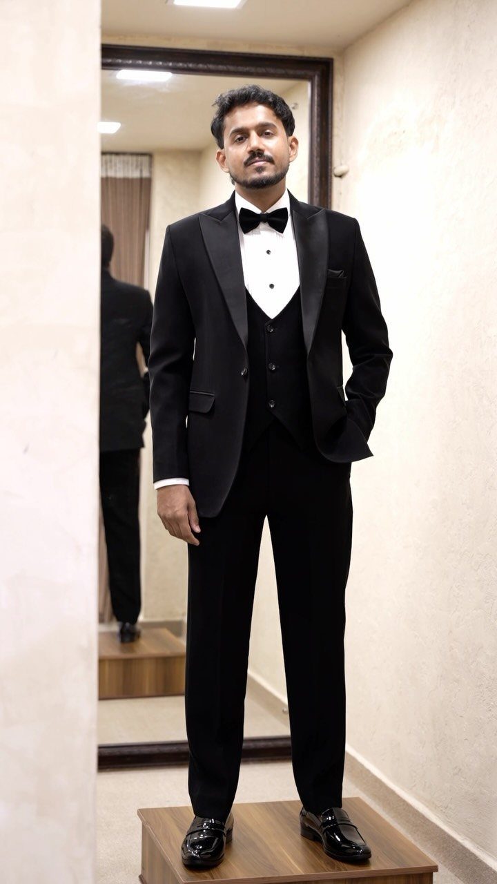 Man posing in a black three-piece tuxedo and bowtie inside a clothing fitting room.