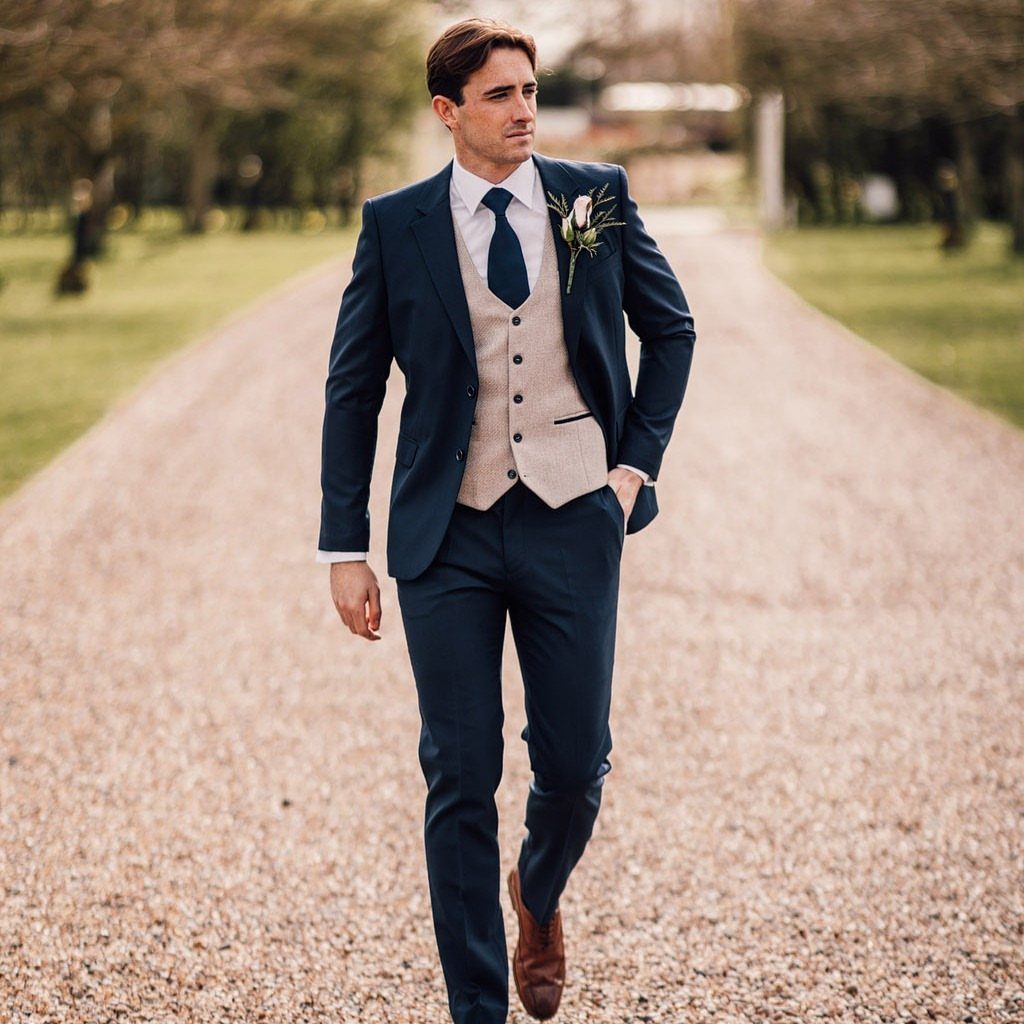 A man in a navy three-piece suit and beige waistcoat walking on a gravel path.